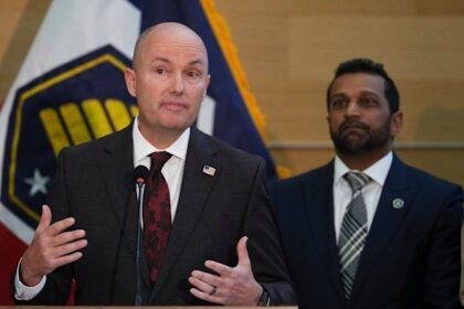 Utah Governor Spencer Cox speaking at a news conference, as FBI Director Kash Patel looks on.