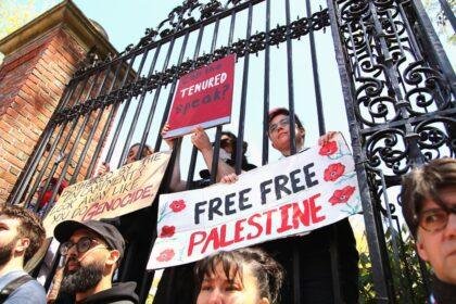 People behind a gate hold signs: "Can the TENURED Speak?", "BOTHERED BY THE ENCAMPMENT? LOOK AWAY, LIKE YOU DO GENOCIDE", and "FREE FREE PALESTINE" with red poppies.